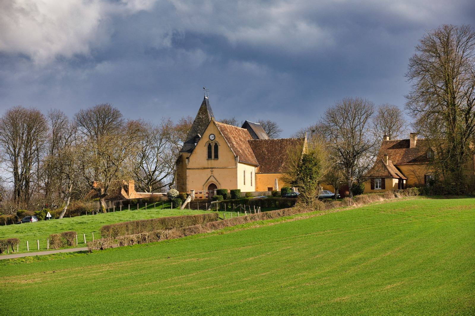 Photographe patrimoine en Sarthe - église de Courcival sur sa butte sous un ciel d'orage, arbres nus en hiver